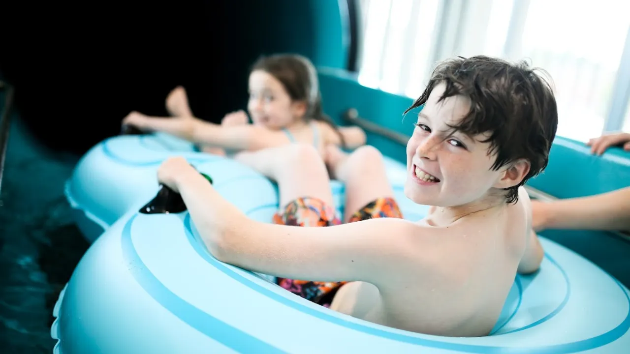 Two children enjoying the inflatable ring slide at Andersonstown Leisure Centre, smiling and seated in blue inner tubes inside the water slide.