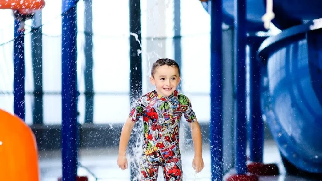 Smiling child running through a water jet in the splash zone at Andersonstown Leisure Centre, wearing a colourful superhero swim outfit.