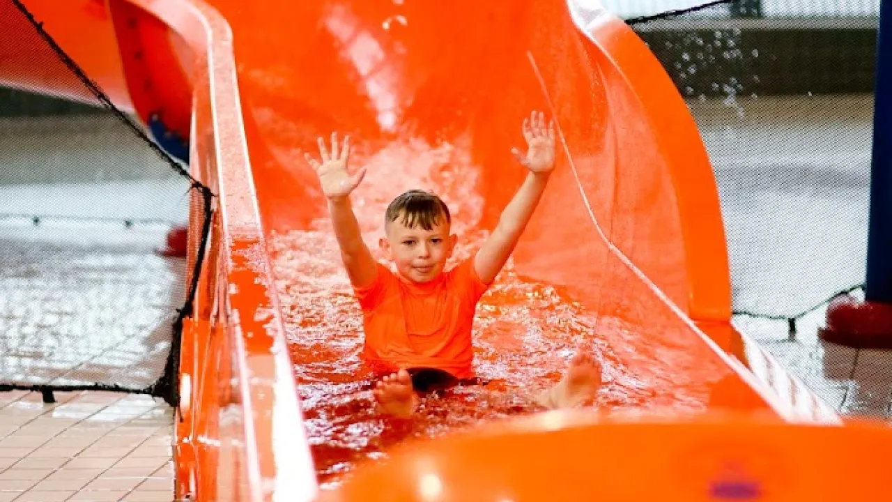 Boy in orange swimwear raising hands while riding down an orange water slide at Andersonstown Leisure Centre’s indoor splash area.