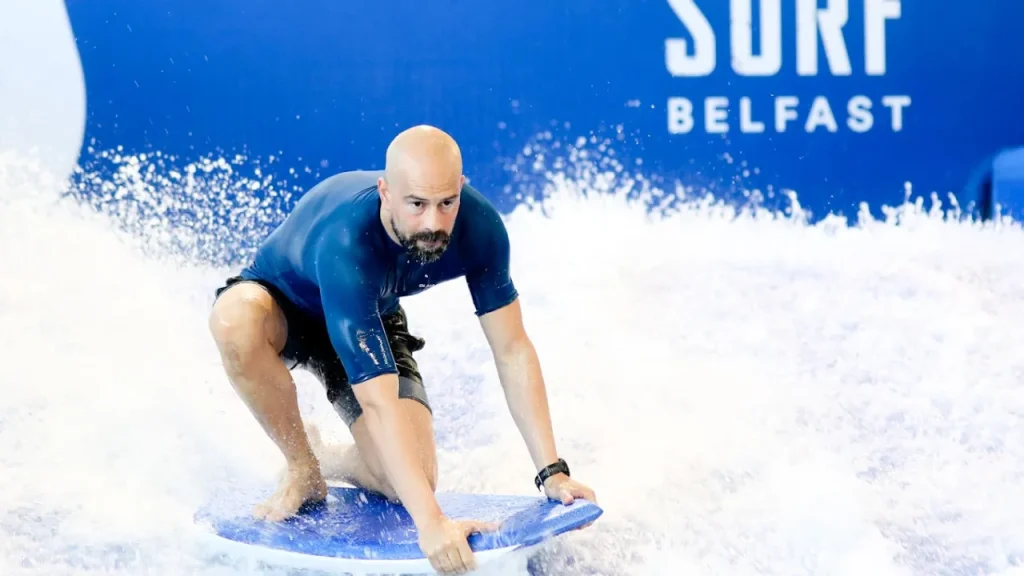Man surfing on the indoor Surf Belfast wave simulator at Andersonstown Leisure Centre, balancing on a blue board in action-packed water flow.