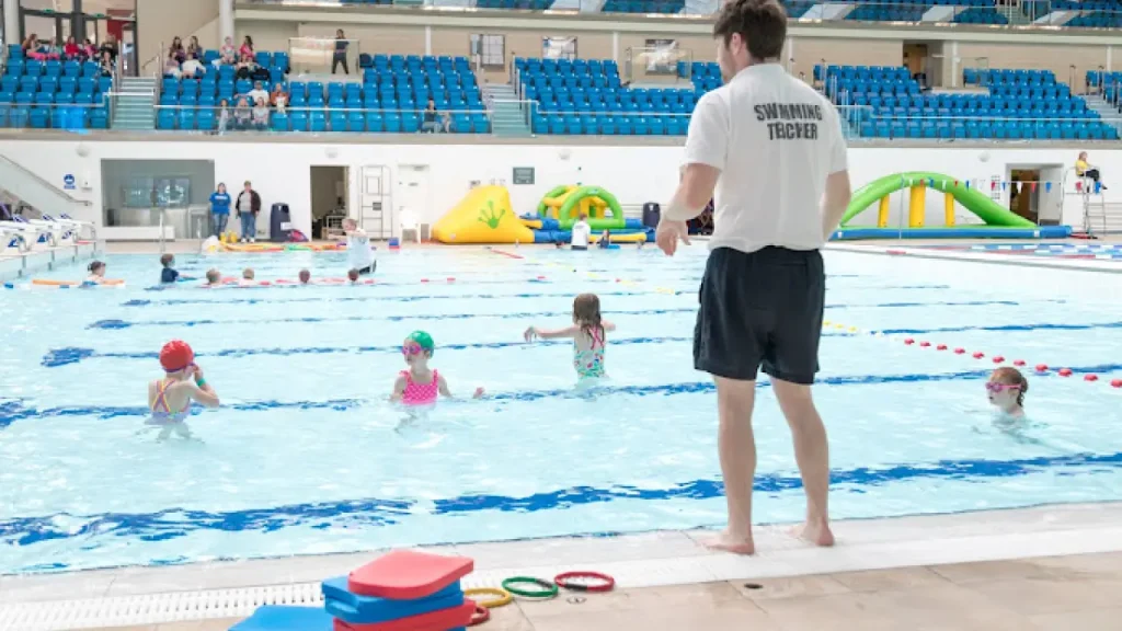 Swimming teacher supervising a children’s swim class at Bangor Aurora Leisure Centre with poolside spectator seating.
