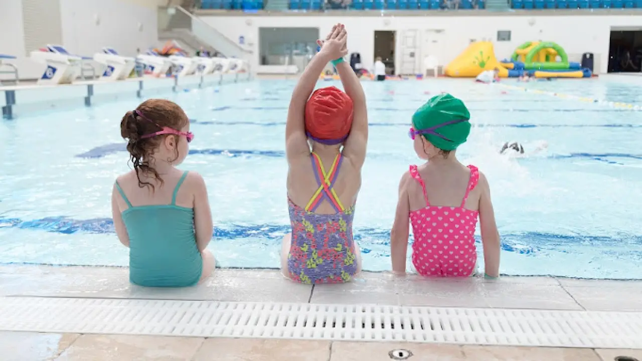 Three young children sitting poolside at Bangor Aurora Leisure Centre, wearing swim caps and preparing for swim activities.
