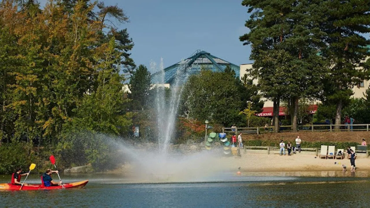 Visitors kayaking on the scenic lake at Center Parcs Elveden Forest, with a water fountain and the iconic Subtropical Swimming Paradise in the background.