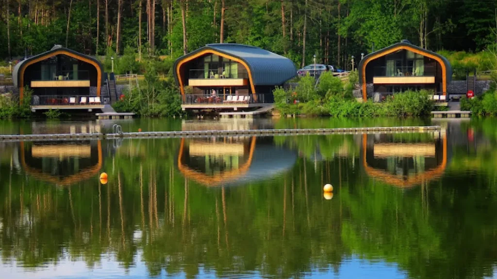 Modern lakeside lodges with curved roofs reflected in the still water at Center Parcs Elveden Forest, surrounded by lush green forest.