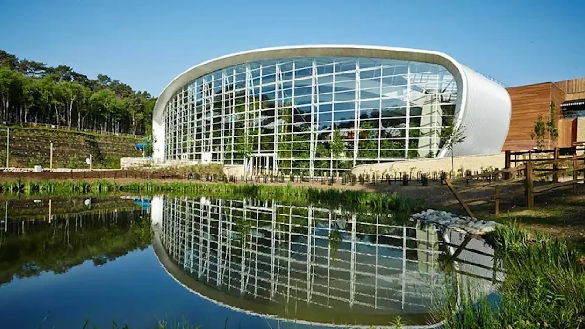 Exterior view of the Subtropical Swimming Paradise at Center Parcs Woburn Forest with a glass façade and pond reflection, surrounded by lush trees.