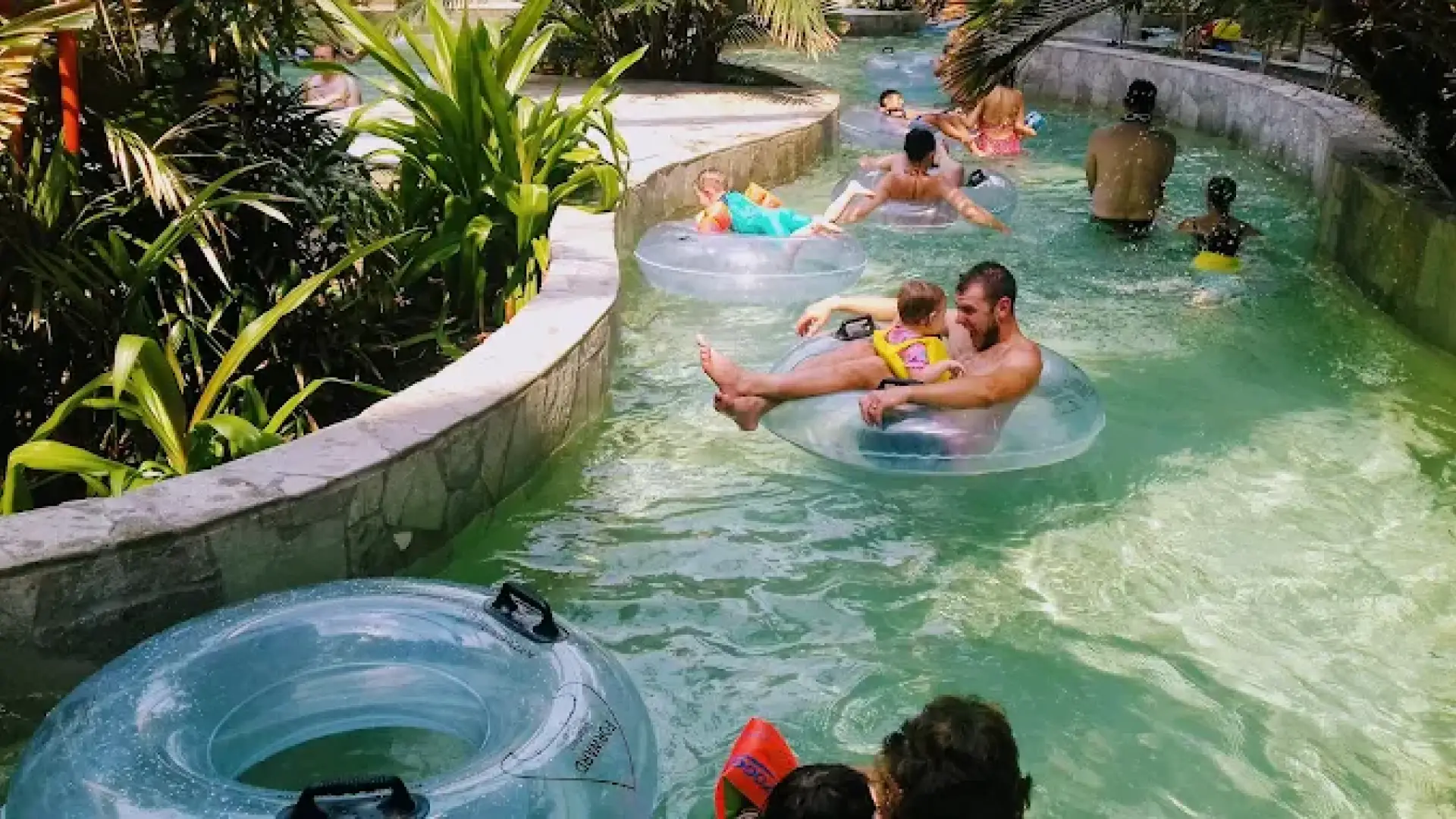 Families floating on rubber rings along the Lazy River at Center Parcs Woburn Forest, with clear water and tropical plants lining the route.