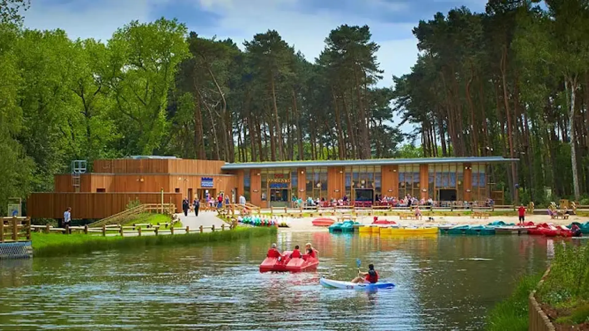 Families boating and kayaking on the lake at Center Parcs Woburn Forest, with the Pancake House and tall pine trees in the background.