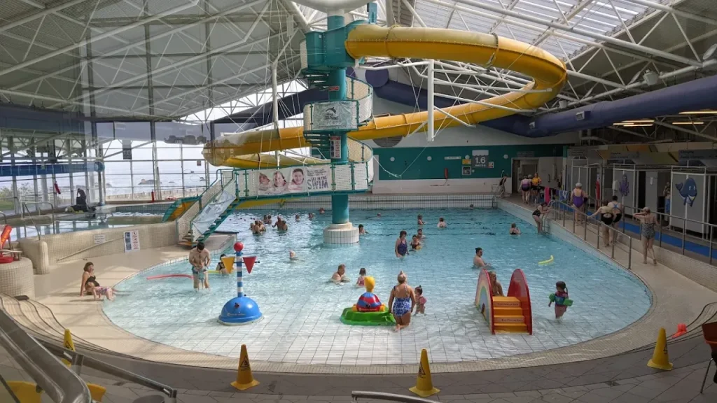 Indoor view of Dunbar Swimming Pool showing families swimming, a yellow flume slide, shallow play areas, and colourful children’s splash toys.