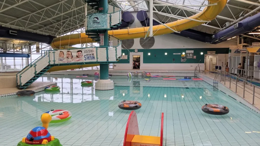 Interior of Dunbar Swimming Pool with yellow flume slide, float rings, inflatable toys, and shallow pool area under a bright glass roof.