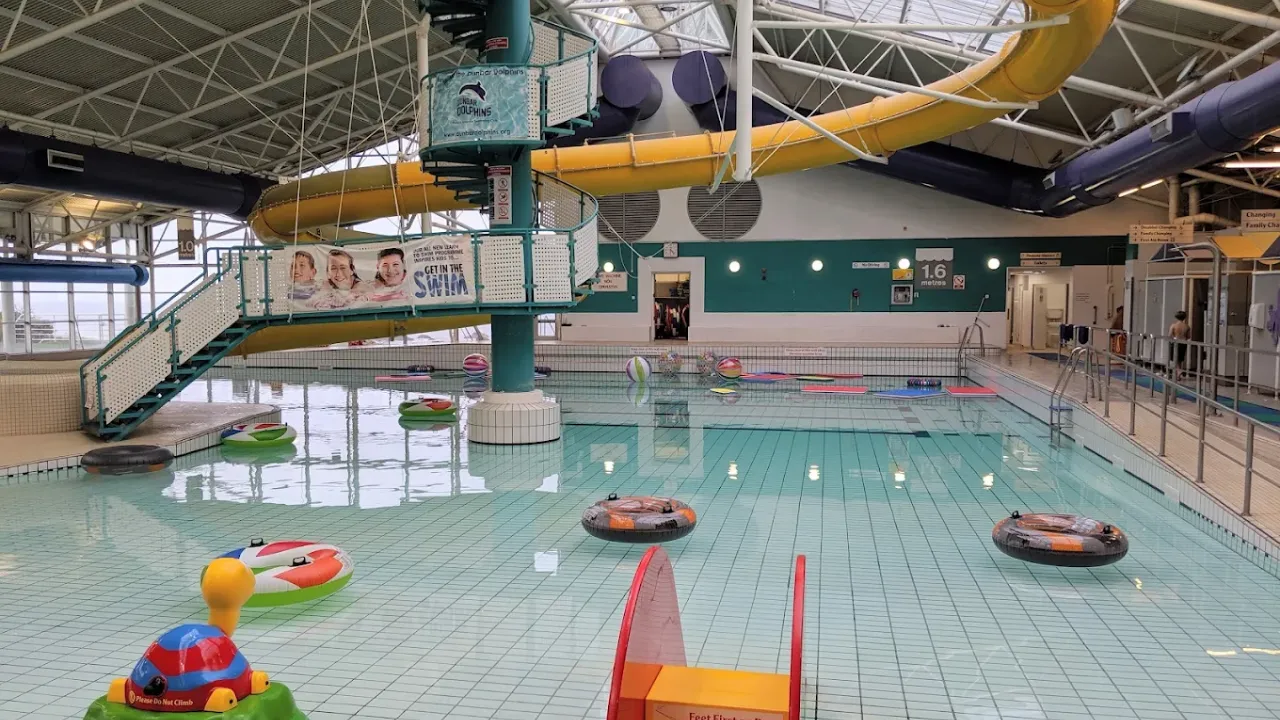 Interior of Dunbar Swimming Pool with yellow flume slide, float rings, inflatable toys, and shallow pool area under a bright glass roof.