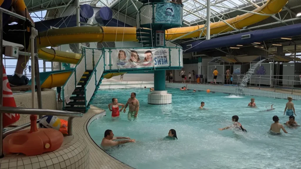 Busy indoor pool scene at Dunbar Swimming Pool with kids splashing, wave machine on, and yellow flume slide winding overhead.