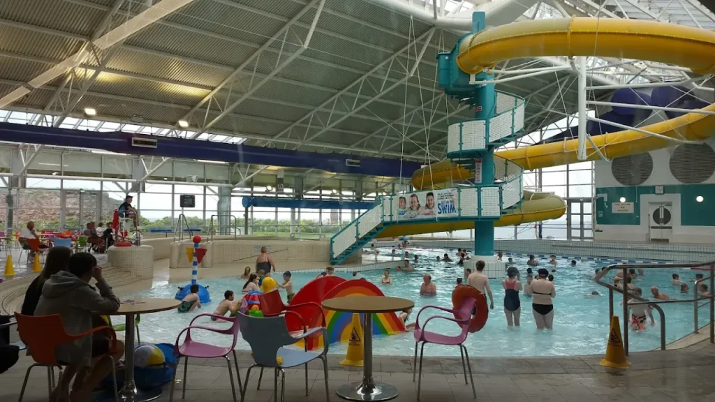 Interior of Dunbar Swimming Pool showing poolside seating, flume slide, rainbow water play features, and families swimming indoors.