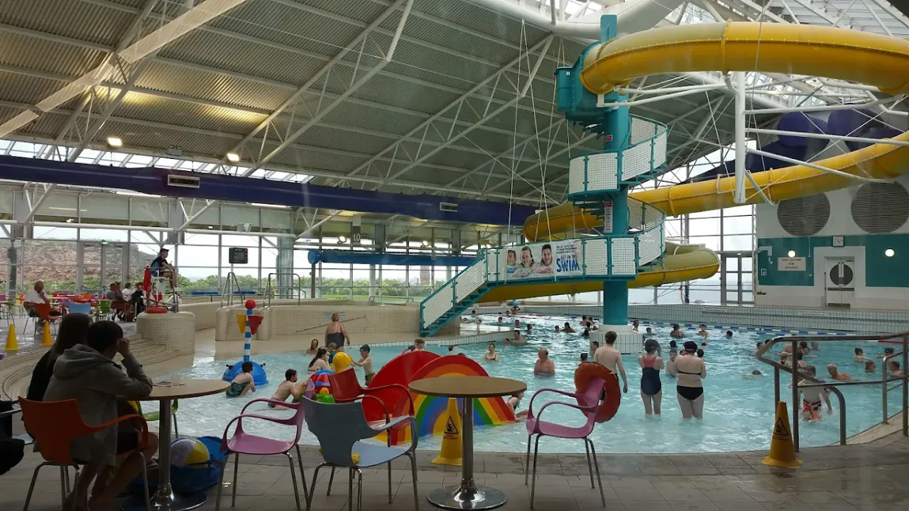 Interior of Dunbar Swimming Pool showing poolside seating, flume slide, rainbow water play features, and families swimming indoors.