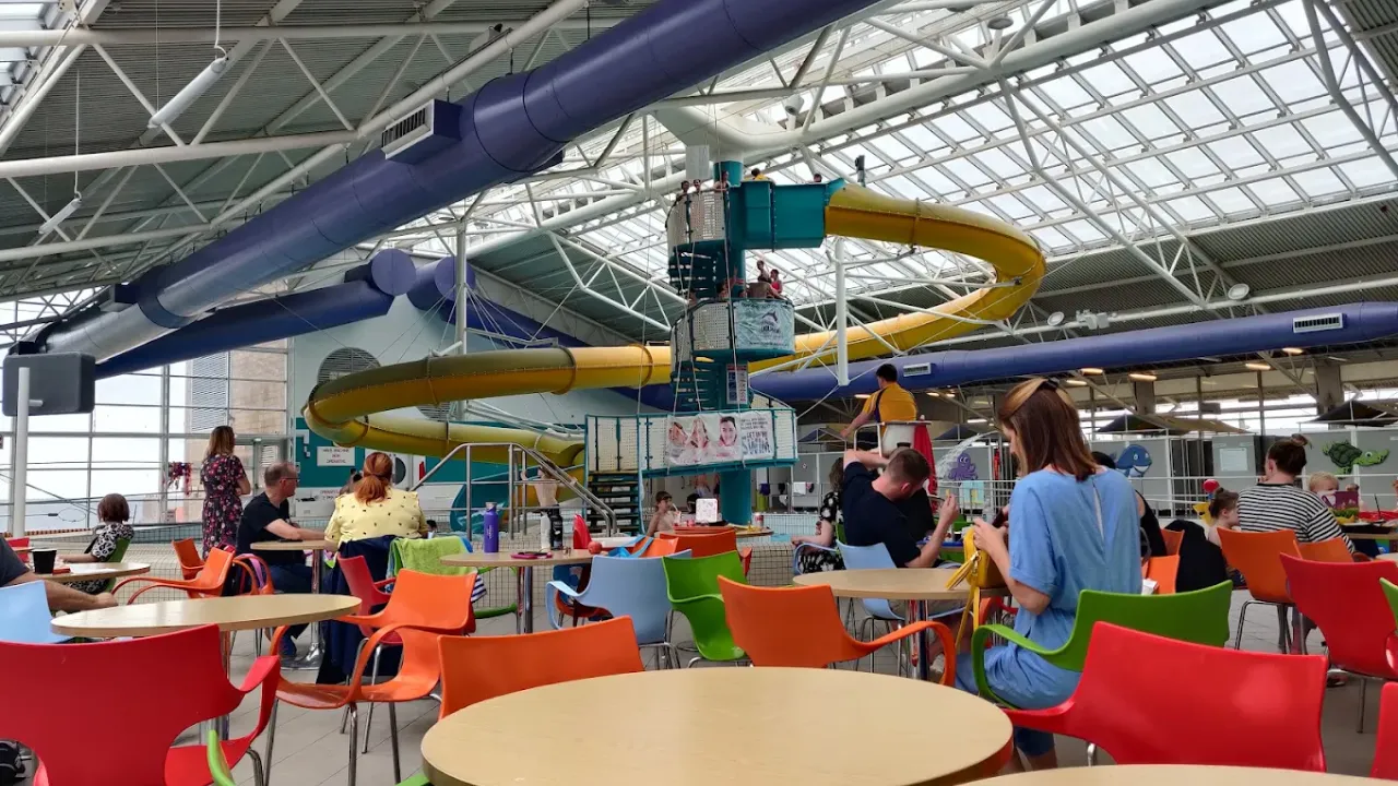 Indoor café seating at Dunbar Swimming Pool with colourful chairs, tables, and a view of the yellow flume and indoor swimming area.