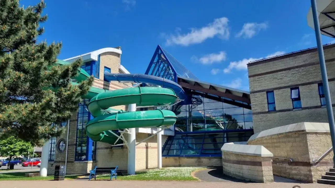Exterior view of Lagan Valley LeisurePlex showing green and blue water slides curving around the building under a bright blue sky.