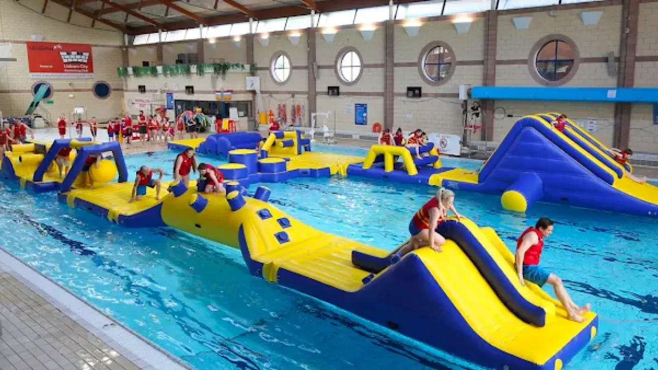 Indoor pool at Lagan Valley LeisurePlex featuring a large yellow and blue inflatable obstacle course with participants climbing and sliding.
