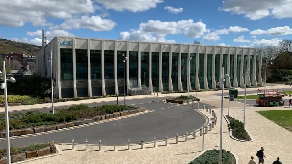 Modern exterior of The LC Swansea leisure centre on a sunny day in South Wales, with landscaped surroundings and pedestrian walkways.