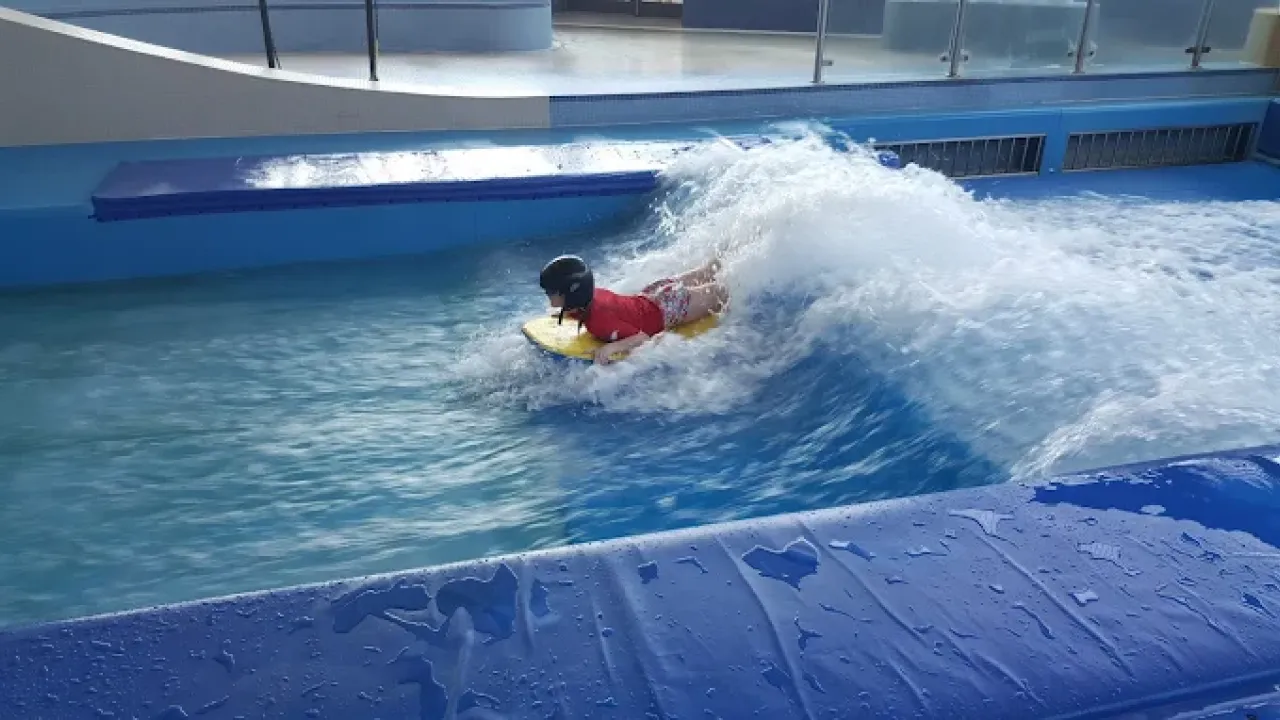Child surfing on the indoor Boardrider wave machine at The LC Swansea, wearing a helmet and riding a bodyboard as water rushes around them.