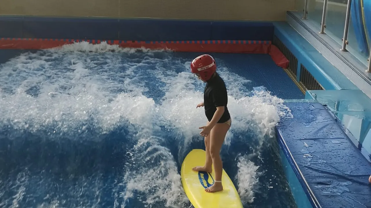 Child standing on a yellow surfboard in helmet on the Boardrider wave simulator at The LC Swansea, riding artificial waves indoors.