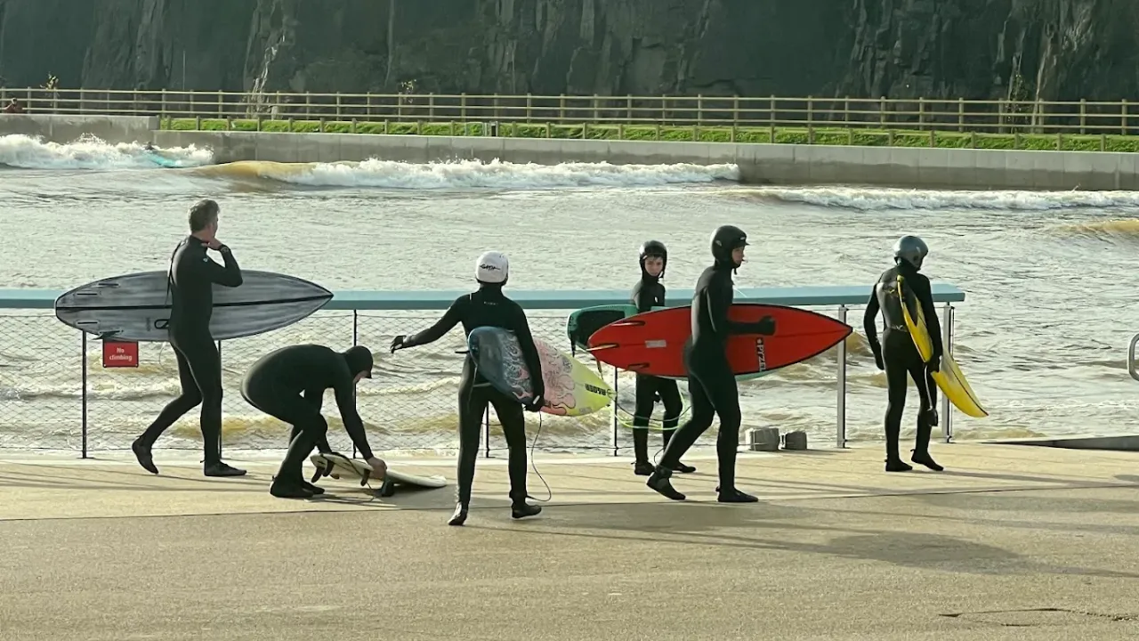 Group of wetsuit-clad surfers with surfboards getting ready beside the artificial wave pool at Lost Shore Surf Resort Edinburgh, with waves rolling in the background.