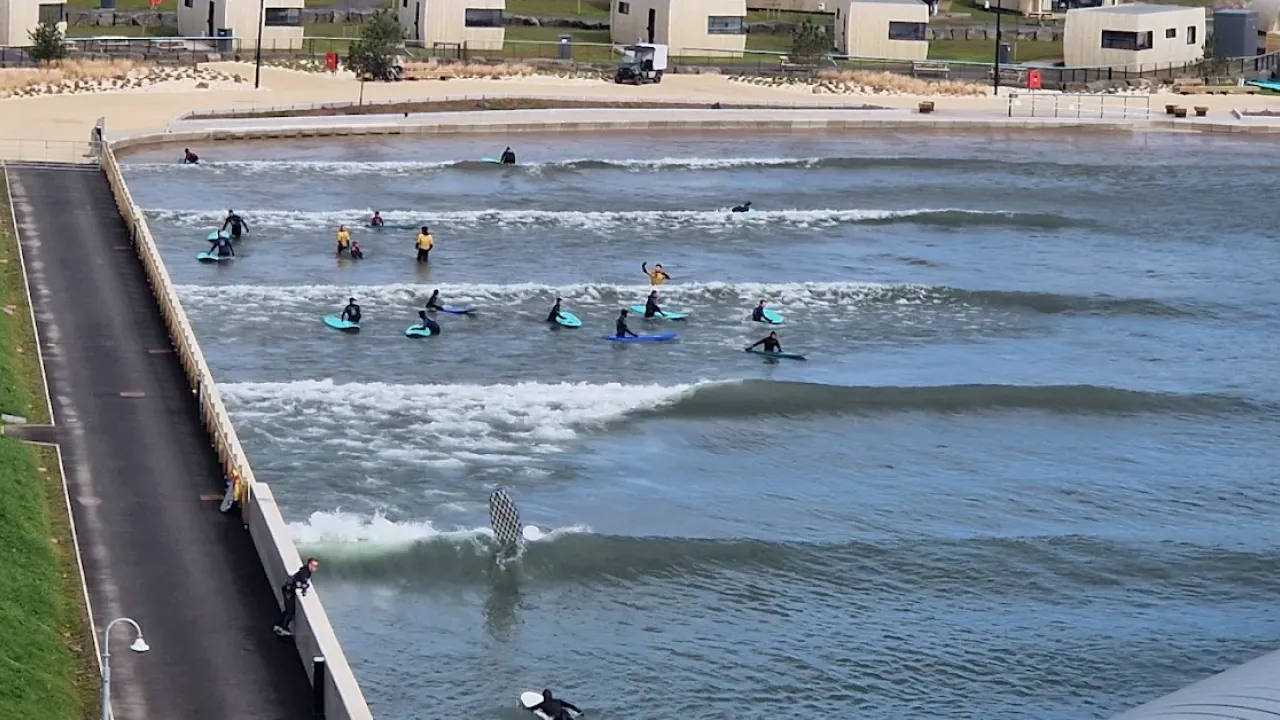 Group of beginner surfers on softboards in the wave pool at Lost Shore Surf Resort Edinburgh, surrounded by modern surf lodges and coaching staff.