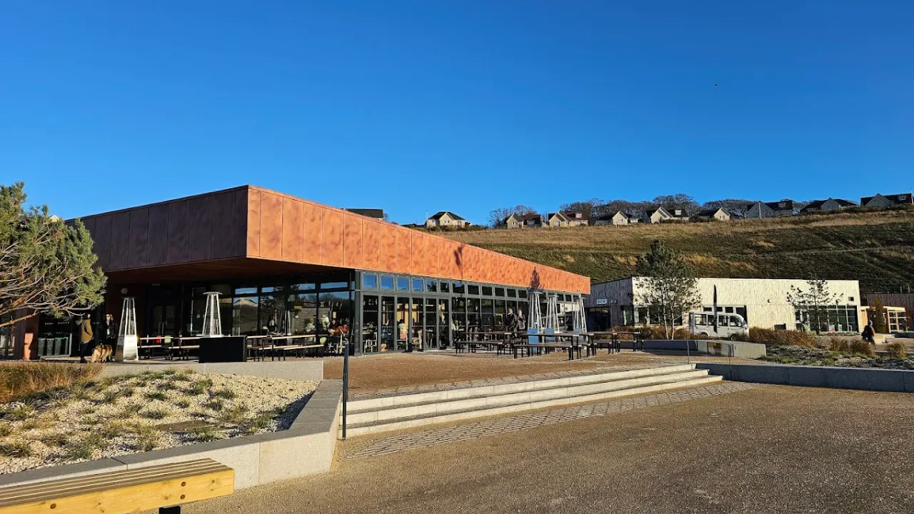 Modern restaurant and café area at Lost Shore Surf Resort Edinburgh with outdoor tables, large glass windows, and hillside backdrop under a clear blue sky.