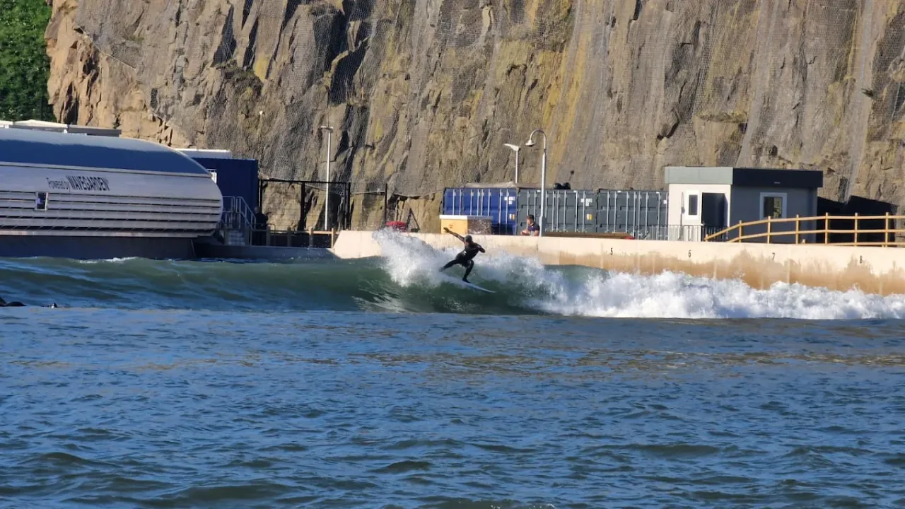 Man surfing a clean artificial wave at Lost Shore Surf Resort in Edinburgh, with Wavegarden technology machinery and steep rock face behind the wave lagoon.