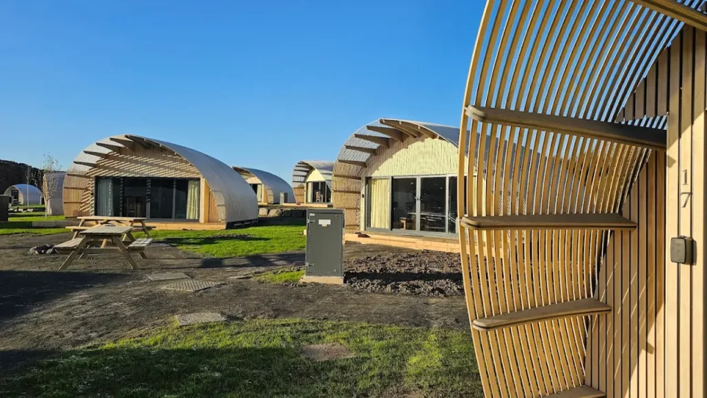 Modern curved Armadilla Pods at Lost Shore Surf Resort Edinburgh with private patios, wooden exteriors, and picnic benches set on grassy plots near the surf lagoon.