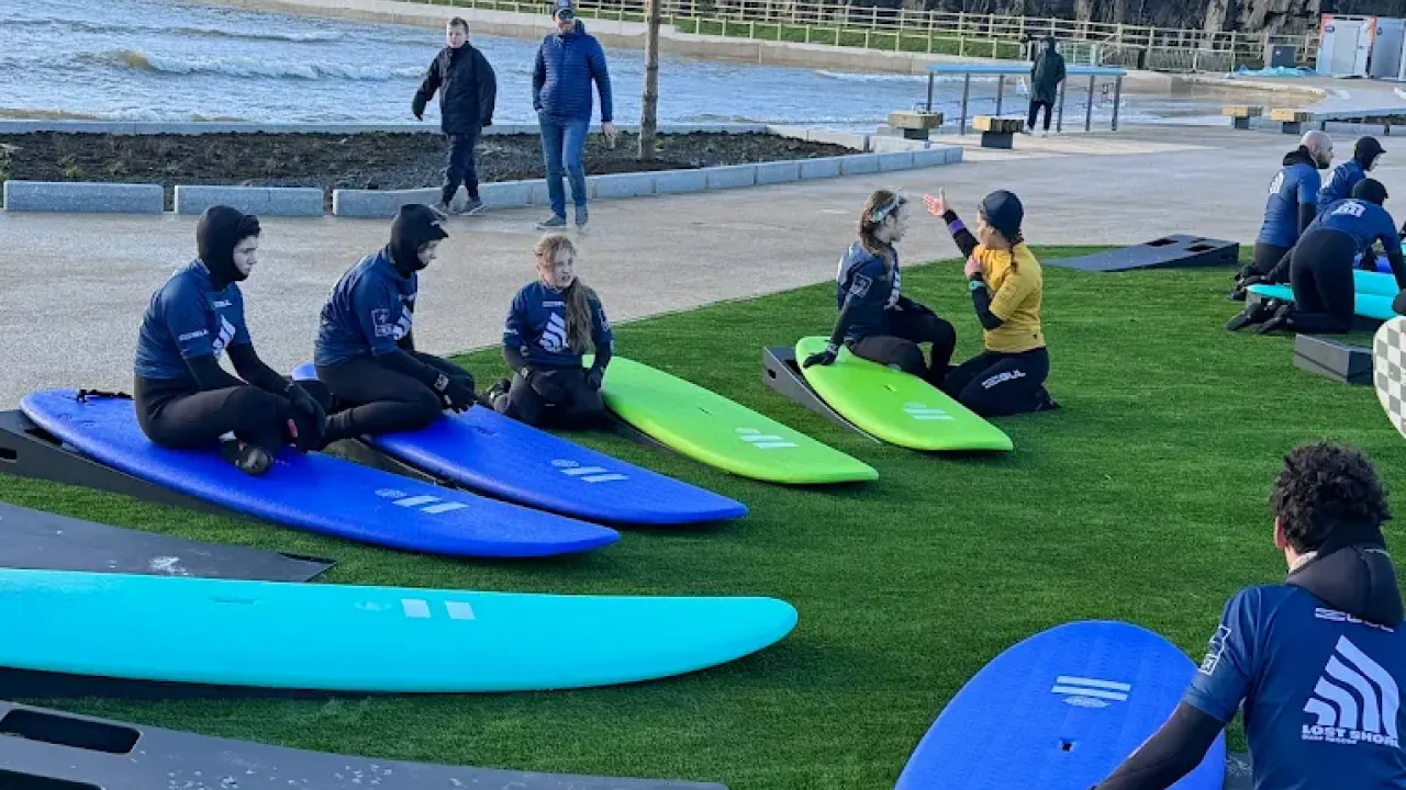 Children in wetsuits sitting on foam surfboards receiving instruction from a surf coach at Lost Shore Surf Resort Edinburgh beside the wave pool.