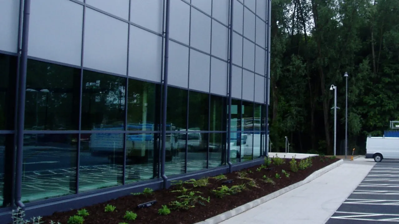 Modern side exterior of Omagh Leisure Complex with glass windows, parking area, and landscaped footpath near forest edge.