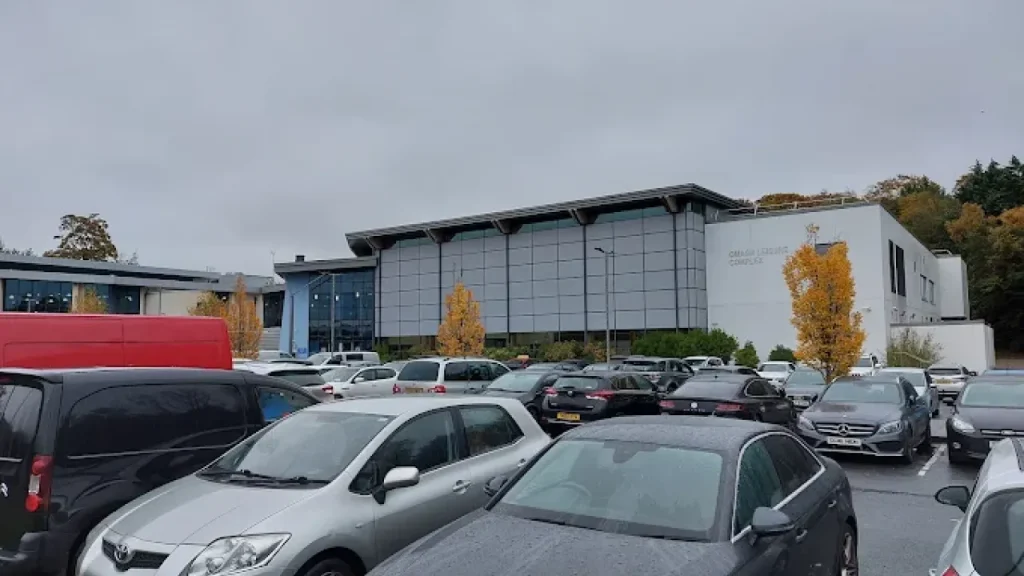 Omagh Leisure Complex with full car park on a cloudy day, featuring the main building and surrounding autumn trees.