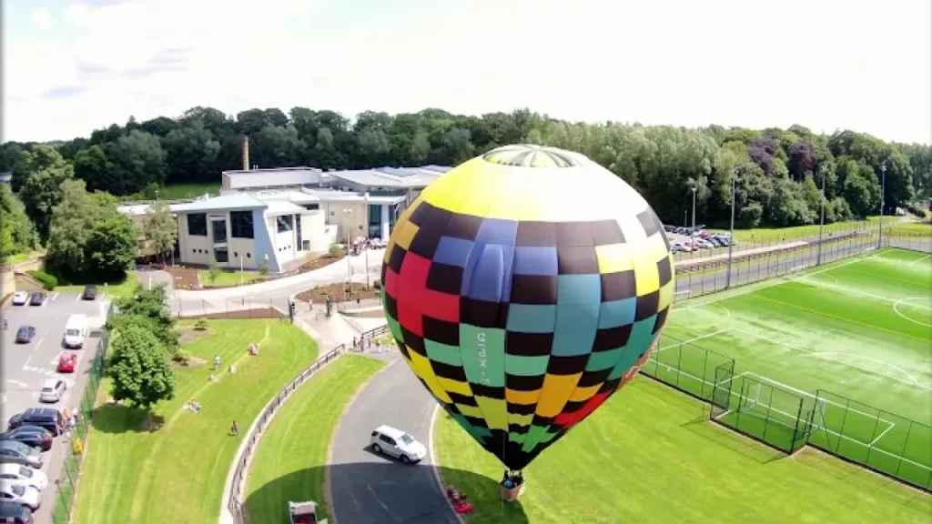 Hot air balloon on green field next to Omagh Leisure Complex and sports pitches during a community event.