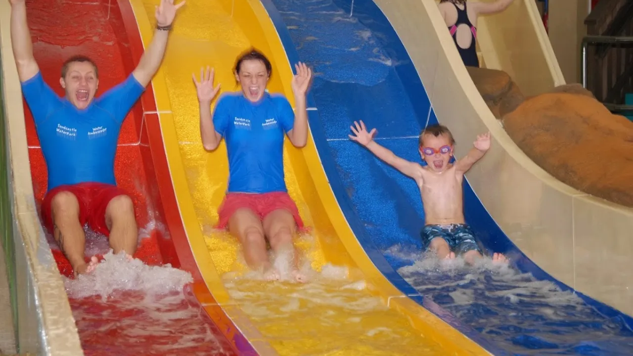 Two adults and a child laughing as they slide down red, yellow, and blue water slides at Sandcastle Waterpark Blackpool.