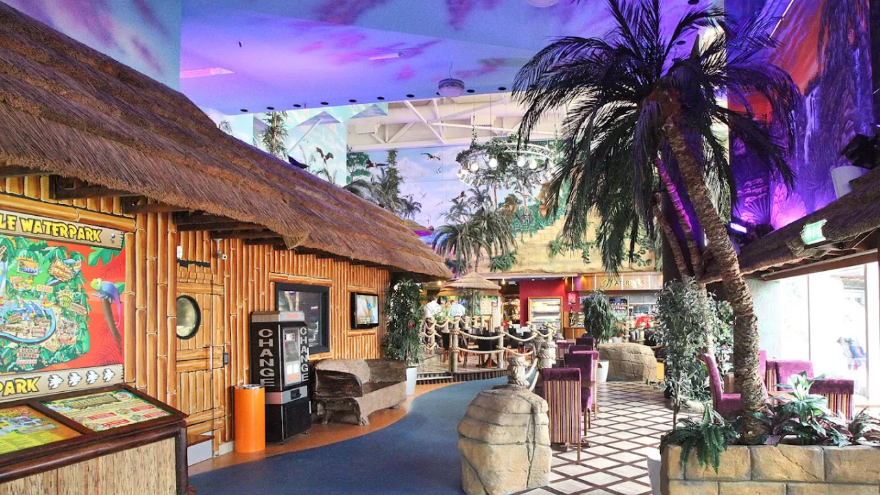Indoor entrance area of Sandcastle Waterpark Blackpool with palm trees, tropical murals, bamboo walls, and seating near Waterfalls Café.