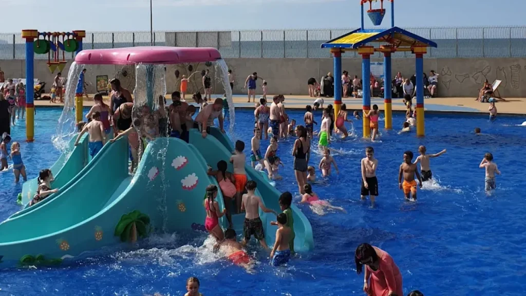 Busy outdoor Splash Pad at SC2 Rhyl featuring children playing on small water slides and under water features during a sunny day.