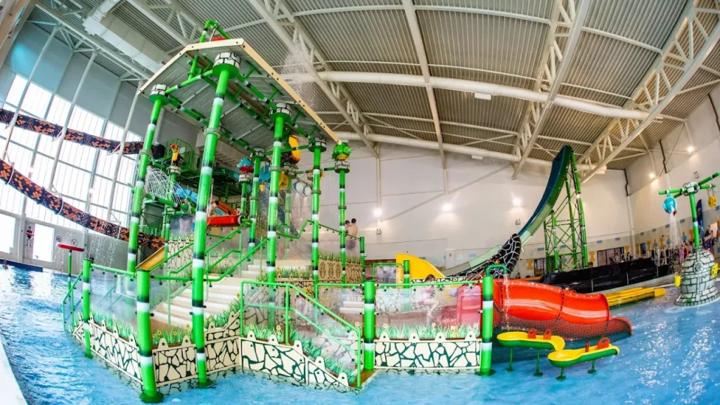 Indoor aquatic play area at SC2 Rhyl with green framework, red and yellow slides, and interactive water features under a high ceiling.