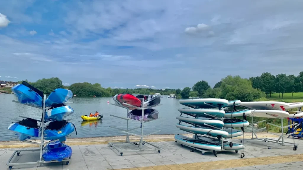 Kayaks, canoes, and paddleboards stacked lakeside at South Lake Leisure Centre with people kayaking in the background