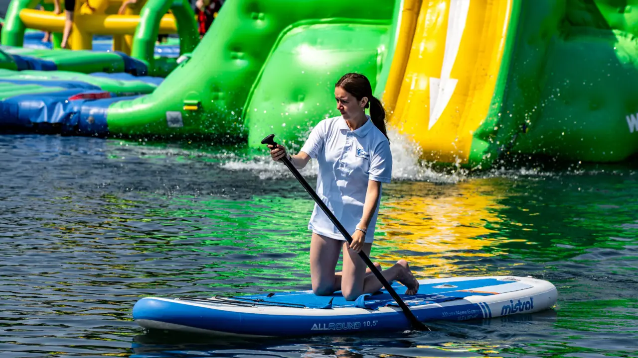 Woman paddleboarding on the lake at Splash Outdoor Waterpark in Northern Ireland, with inflatable slides and obstacle course behind.