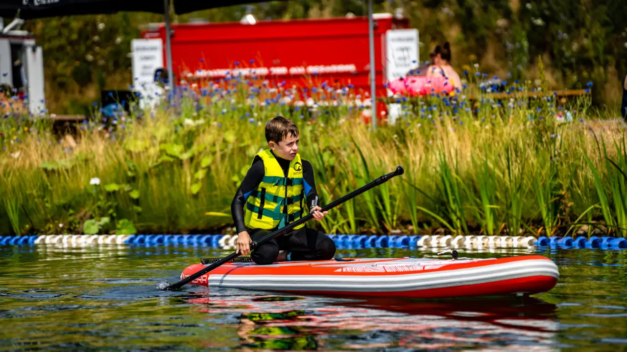 Child paddleboarding on the lake at Splash Outdoor Waterpark in Northern Ireland, wearing a yellow safety vest with grassy shoreline in background.