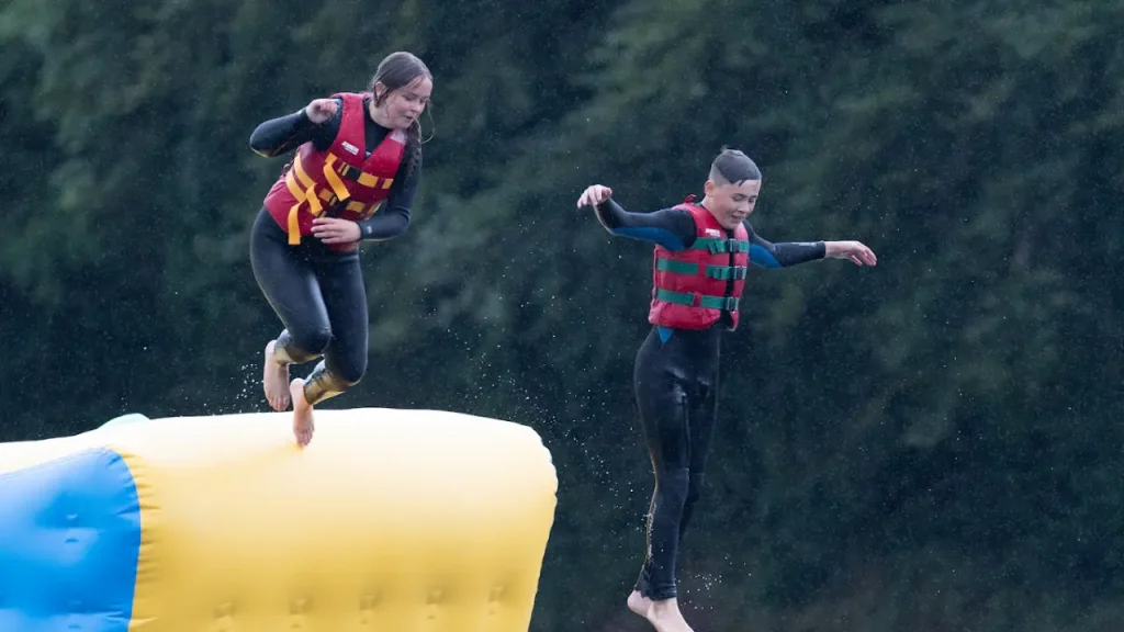 A girl and boy in red life jackets jumping off a large yellow inflatable platform at The Lake Kilrea Waterpark, with water splashing and trees in the background.