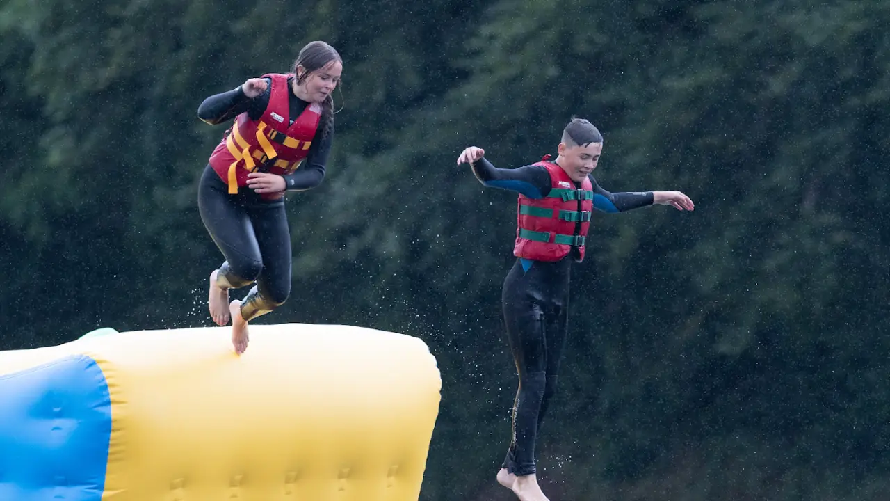 A girl and boy in red life jackets jumping off a large yellow inflatable platform at The Lake Kilrea Waterpark, with water splashing and trees in the background.