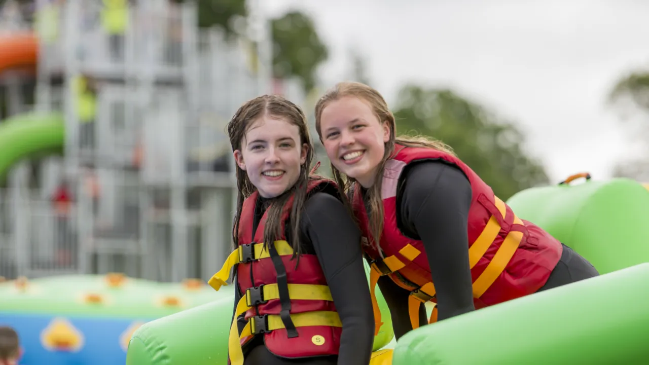 Two girls in wetsuits and red buoyancy aids on a green inflatable at the Lake Kilrea waterpark, Kilrea, Northern Ireland.