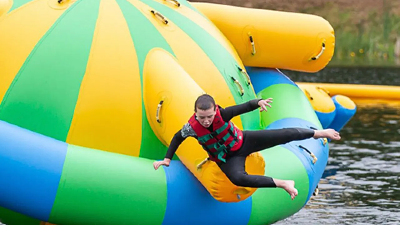 Child in wetsuit and red buoyancy aid sliding off a green-yellow inflatable on the Lake Kilrea waterpark’s floating course in Kilrea, County Londonderry.