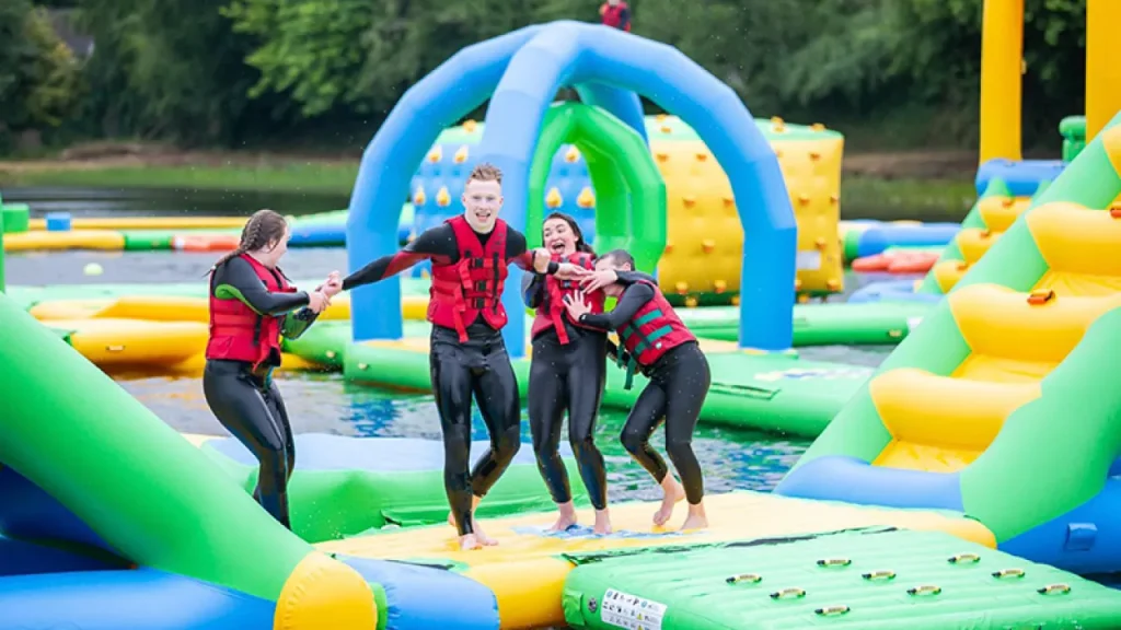 Four visitors in red buoyancy aids running over green and yellow floating platforms on the Lake Kilrea waterpark’s inflatable course, with blue arches and climb walls behind in Kilrea, County Londonderry.