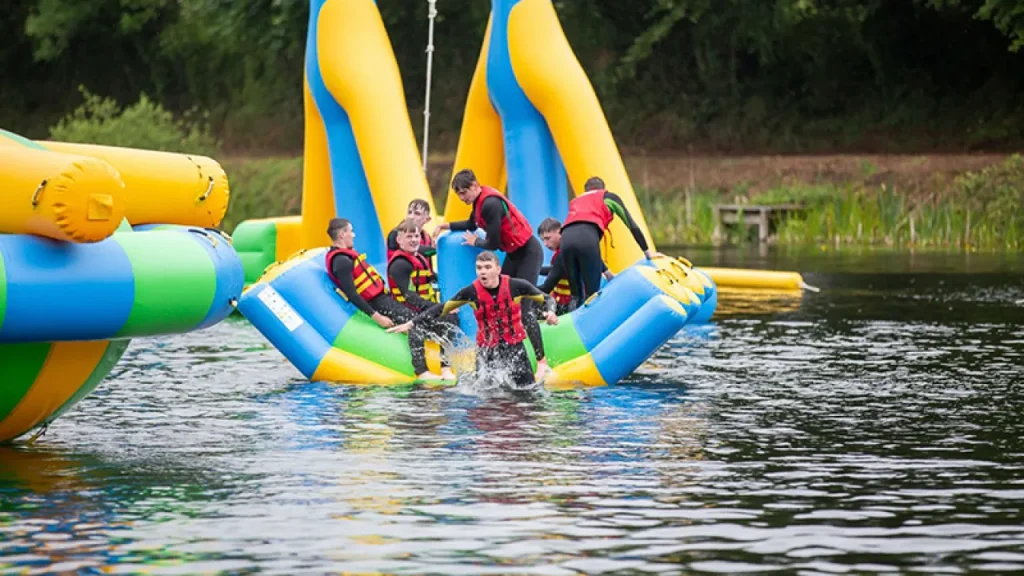 Group of teenagers in red buoyancy aids tipping from a blue-green inflatable into Lake Kilrea, with tall blue-and-yellow slide towers in the background, Kilrea, County Londonderry.