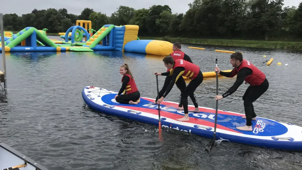 Group on a giant stand-up paddleboard (Big SUP) wearing red buoyancy aids near the Lake Kilrea waterpark’s green-and-yellow inflatables in Kilrea, County Londonderry.