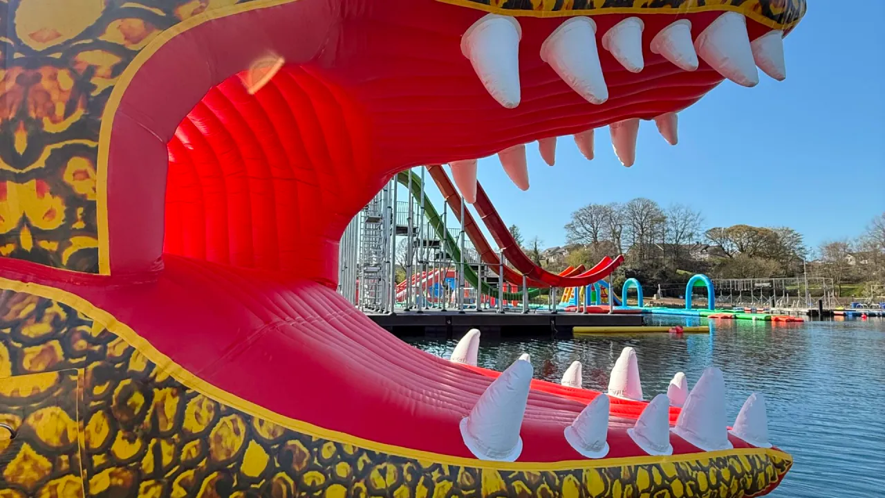 Close-up of a red inflatable mouth with white teeth at The Lake Kilrea, looking towards multi-lane red and green slides and blue-green inflatable arches on the lake in Kilrea, County Londonderry.