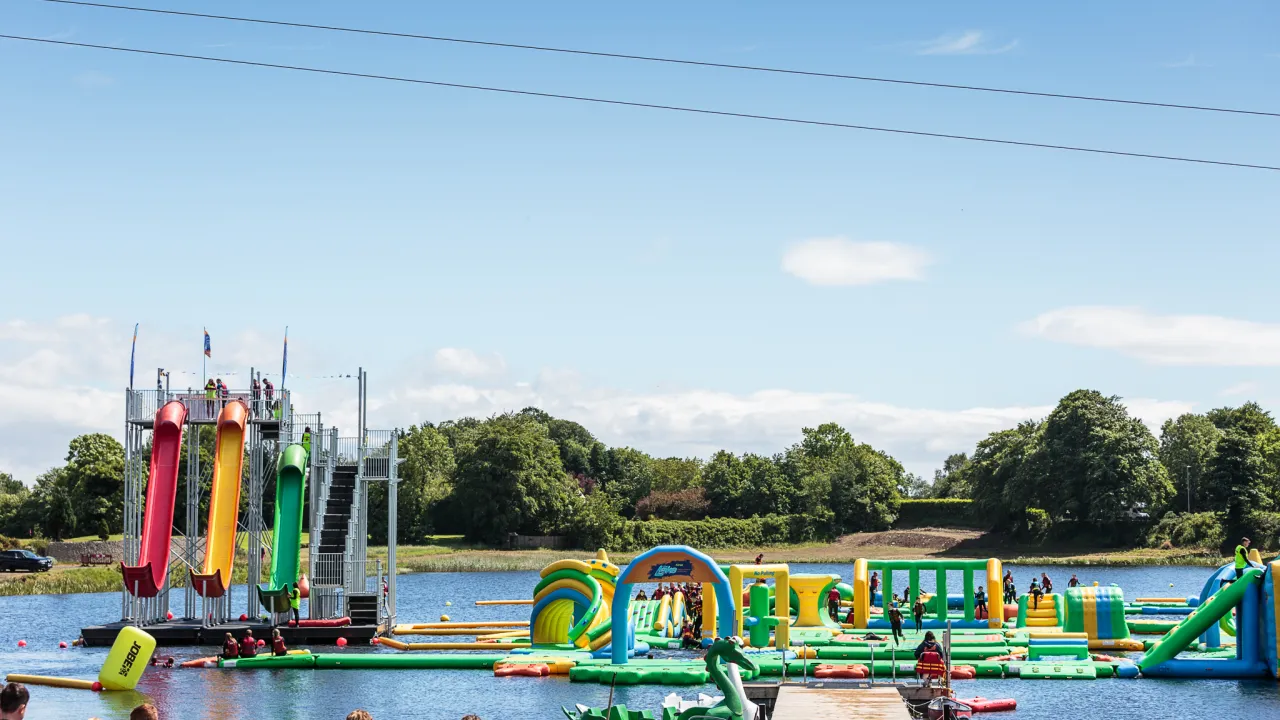 Wide view of The Lake Kilrea waterpark showing red, yellow and green multi-lane slide tower and green-yellow inflatables on the lake at Kilrea, County Londonderry.