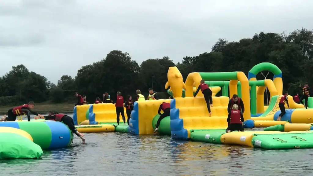 Visitors in life jackets climbing and sliding on colourful yellow and green inflatable obstacles at The Lake Kilrea Waterpark in Northern Ireland.