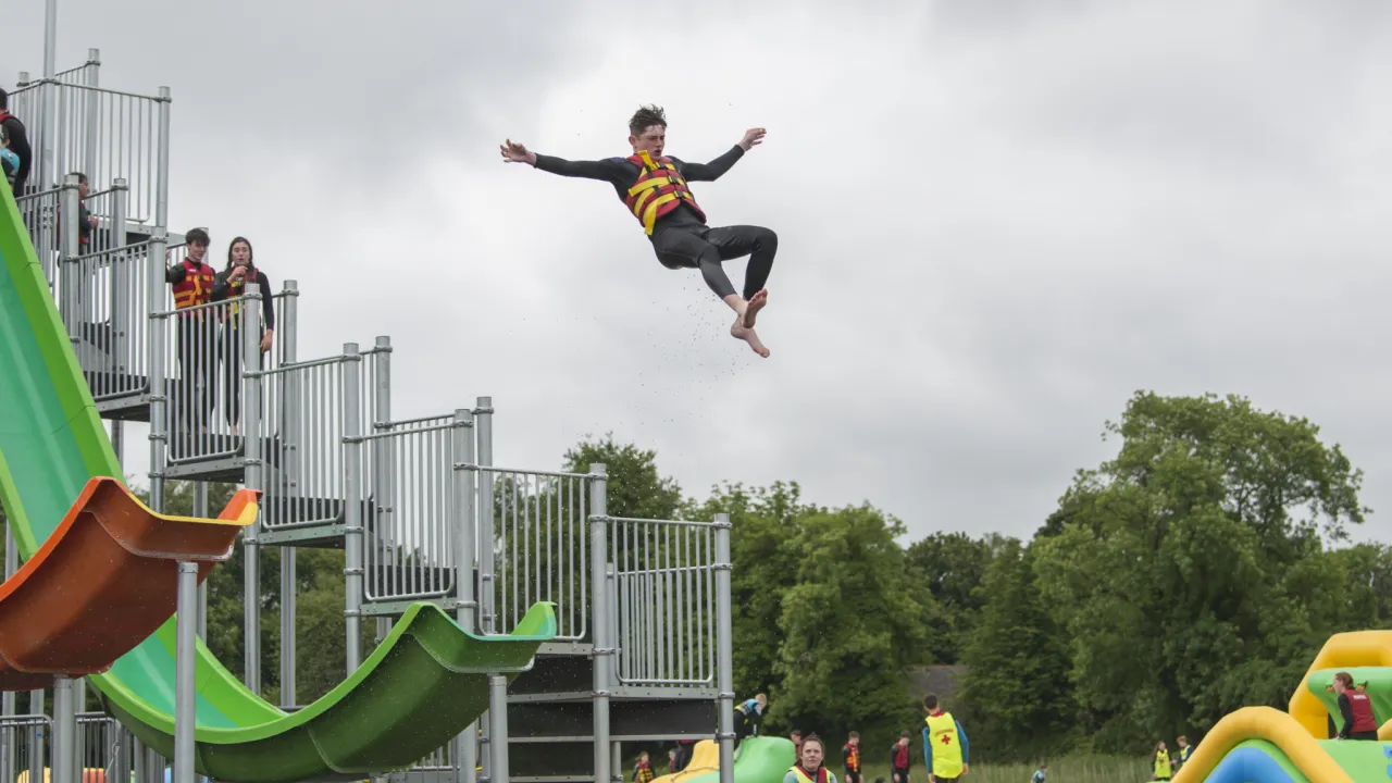 Participant in a wetsuit and red–yellow buoyancy aid airborne from a green launch slide at The Lake Kilrea waterpark, with the slide tower and spectators behind in Kilrea, County Londonderry.